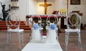 Przezroczyste krzesła ozdobione niebieskimi kwiatami i białą tkaniną, ustawione w kościele przed ołtarzem.