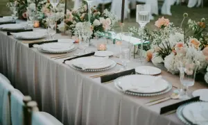 Elegancka dekoracja sali weselnej, gdzie cena idzie w parze z pięknem. Stoły nakryte jasnym obrusem, ozdobione kwiatami i świecami.