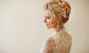 Eleganckie upięcia okolicznościowe na blond włosach ozdobione perłami i kryształami. Delikatny makijaż i koronkowa suknia podkreślają urodę modelki.