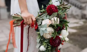 Dłoń panny młodej trzyma bukiet z bordowymi daliami i kremowymi różami, ozdobiony czerwonymi wstążkami. Idealne kwiaty sezonowe na wrzesień.