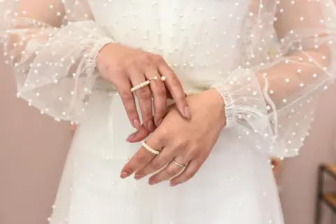 Dłonie panny młodej ozdobione pierścionkami z perłami, na tle sukni ślubnej z delikatnymi koralikami. Perły na ślubie dodają elegancji.