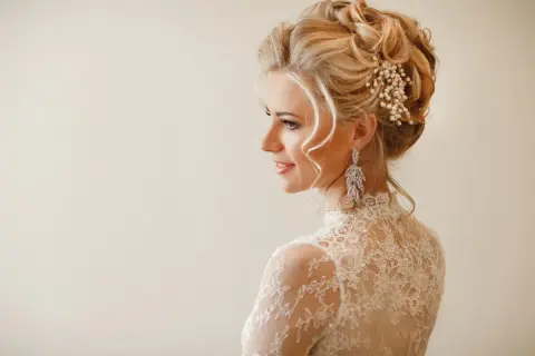 Eleganckie upięcia okolicznościowe na blond włosach ozdobione perłami i kryształami. Delikatny makijaż i koronkowa suknia podkreślają urodę modelki.