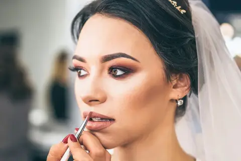 Panna młoda z welonem i biżuterią otrzymuje makijaż ślubny glamour. Usta podkreślone błyszczykiem, oczy smoky eye.