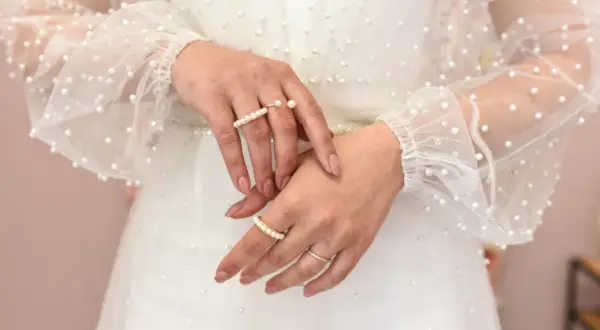 Dłonie panny młodej ozdobione pierścionkami z perłami, na tle sukni ślubnej z delikatnymi koralikami. Perły na ślubie dodają elegancji.