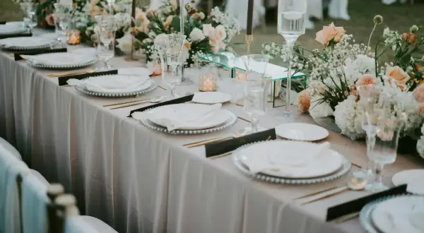 Elegancka dekoracja sali weselnej, gdzie cena idzie w parze z pięknem. Stoły nakryte jasnym obrusem, ozdobione kwiatami i świecami.