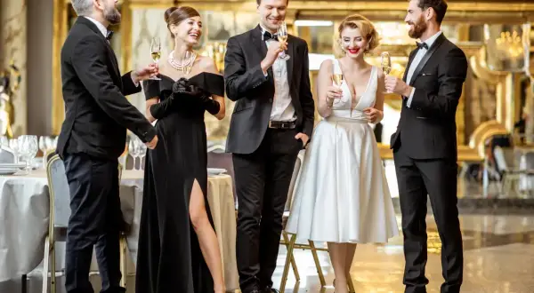 Eleganckie kobiety w sukniach wieczorowych i mężczyźni w garniturach, zgodnie z dress codem black tie, wznoszą toasty.