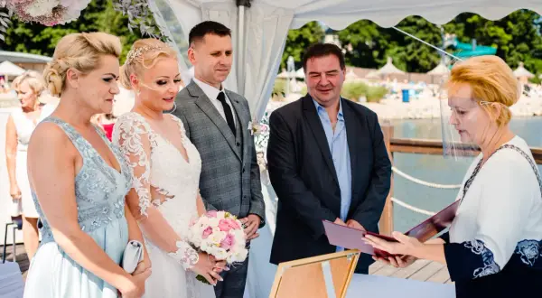 Para ślubna, świadkowie i urzędniczka z przyłbicą podczas ceremonii ślubnej nad jeziorem.
