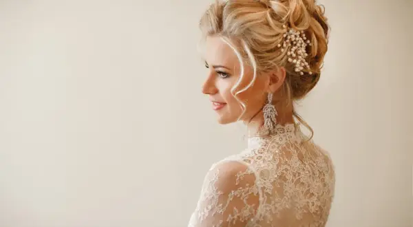 Eleganckie upięcia okolicznościowe na blond włosach ozdobione perłami i kryształami. Delikatny makijaż i koronkowa suknia podkreślają urodę modelki.
