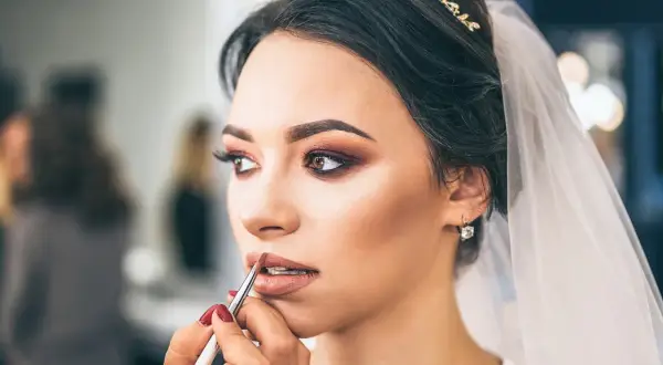 Panna młoda z welonem i biżuterią otrzymuje makijaż ślubny glamour. Usta podkreślone błyszczykiem, oczy smoky eye.