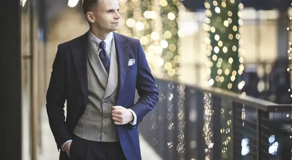 Elegancki mężczyzna w granatowym garniturze i kamizelce, gotowy na wieczór w stylu cocktail attire, na tle migoczących świateł.