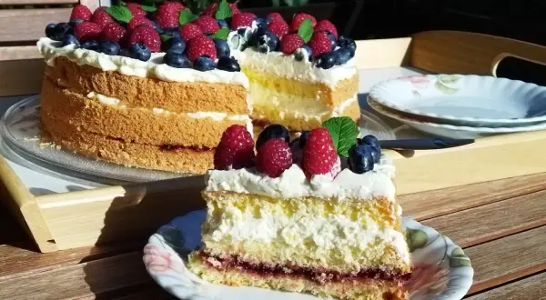 Prosty tort biszkoptowy z kremem i owocami leśnymi, udekorowany malinami i borówkami.