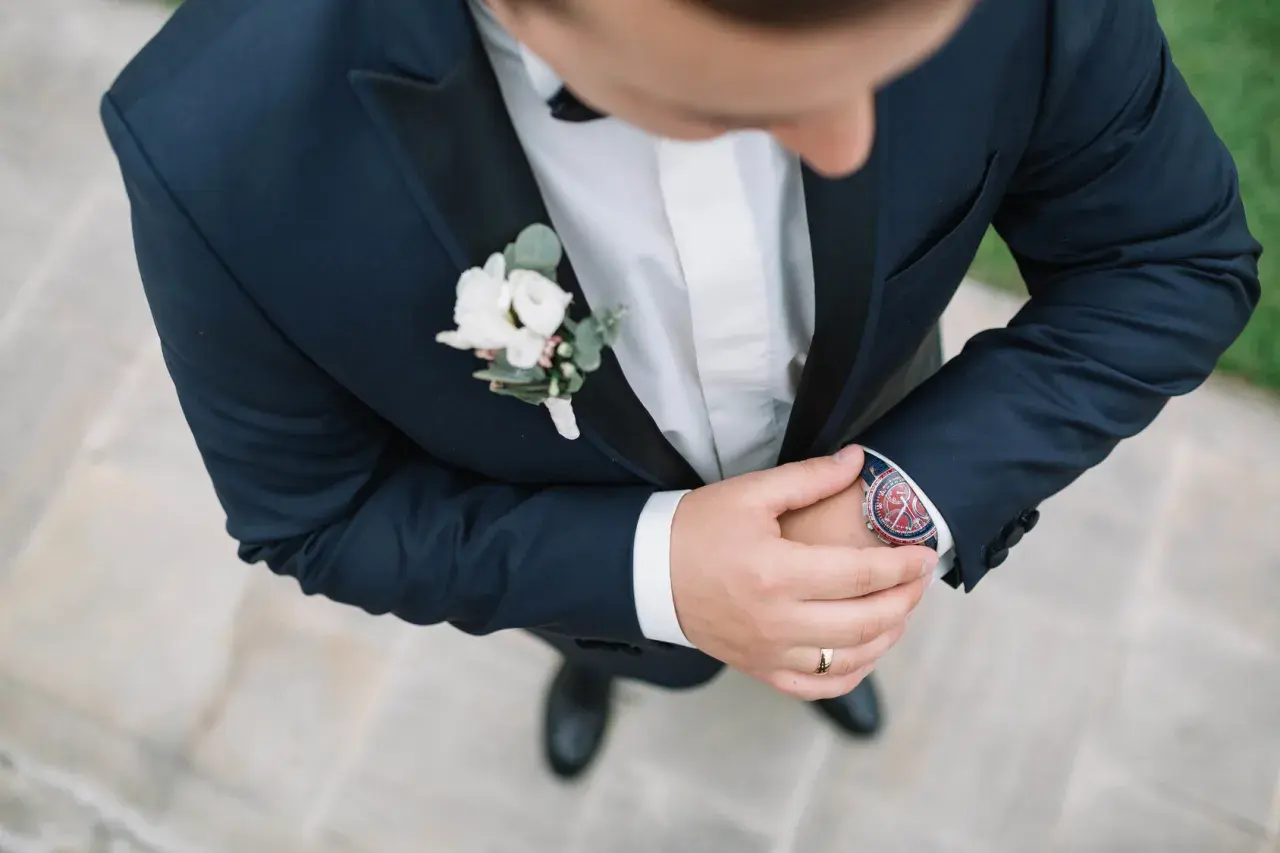 Elegancka butonierka dla pana młodego i świadka, ozdobiona białymi kwiatami i zielonymi liśćmi, dodaje szyku granatowemu garniturowi.