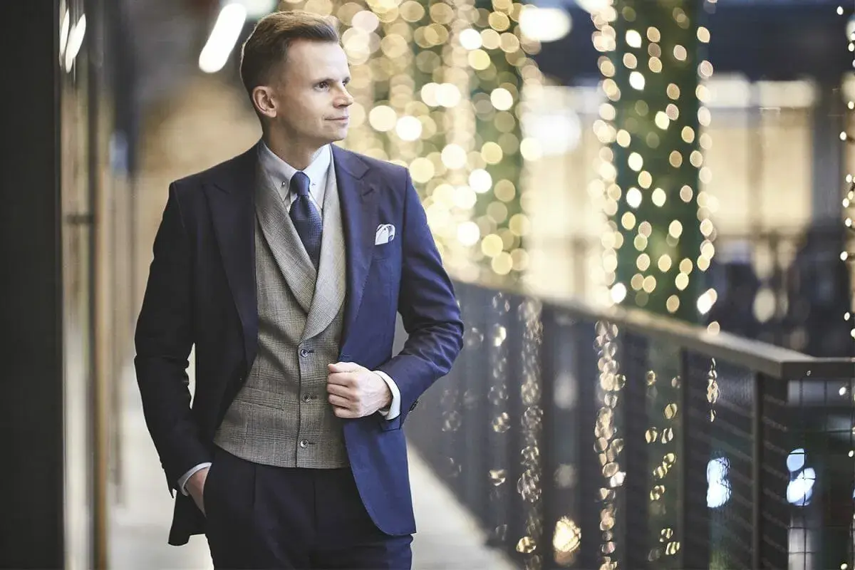 Elegancki mężczyzna w granatowym garniturze i kamizelce, gotowy na wieczór w stylu cocktail attire, na tle migoczących świateł.