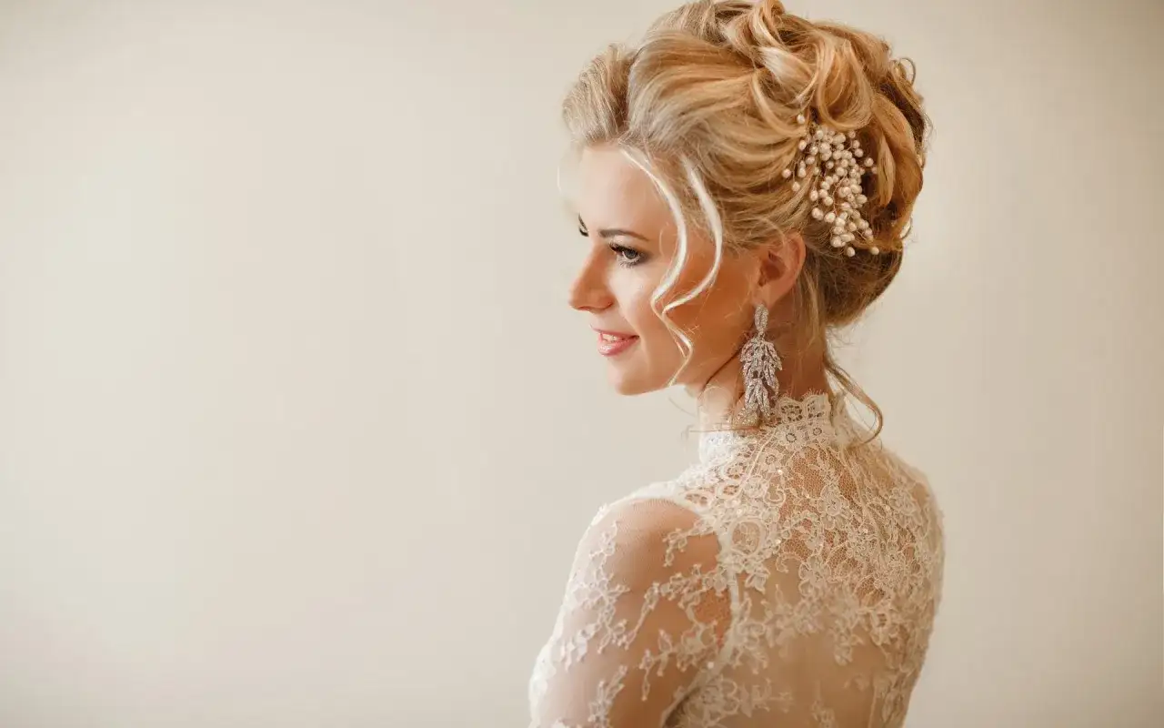 Eleganckie upięcia okolicznościowe na blond włosach ozdobione perłami i kryształami. Delikatny makijaż i koronkowa suknia podkreślają urodę modelki.