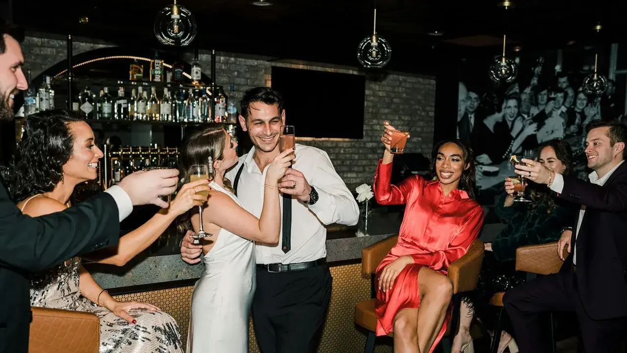 Grupa przyjaciół w eleganckich strojach cocktailowych wznosi toasty w barze.