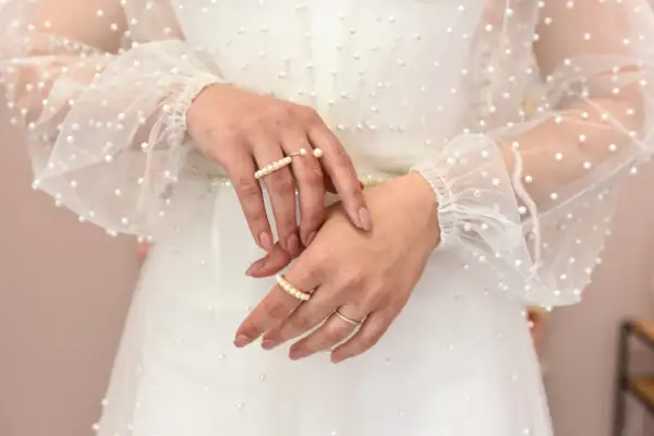 Dłonie panny młodej ozdobione pierścionkami z perłami, na tle sukni ślubnej z delikatnymi koralikami. Perły na ślubie dodają elegancji.