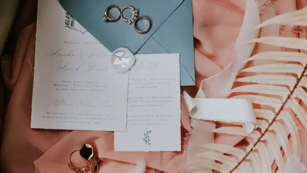 Eleganckie zaproszenia ślubne, obrączki i ozdobne liście na różowym tle.