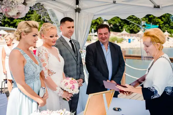 Para ślubna, świadkowie i urzędniczka z przyłbicą podczas ceremonii ślubnej nad jeziorem.