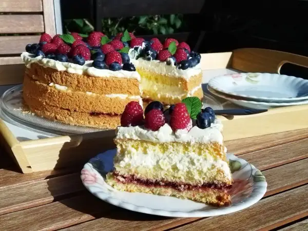 Prosty tort biszkoptowy z kremem i owocami leśnymi, udekorowany malinami i borówkami.